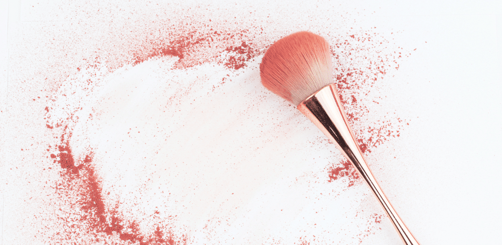 Image of a makeup brush on a white surface surrounded by blusher powder.