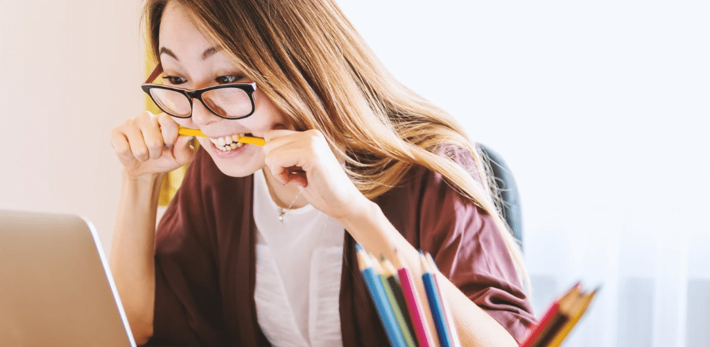 Image of lady sat at a desk with a pencil clenched between her teeth, looking at a laptop screen.