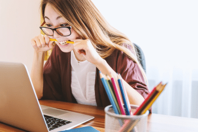 Image of lady sat at a desk with a pencil clenched between her teeth, looking at a laptop screen.